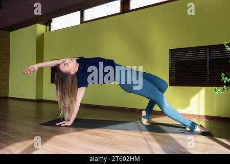 Young attractive brown-haired woman practicing yoga, performing Kamatkarasana exercise, dancing dog pose, training in sportswear standing on a mat in Stock Photo