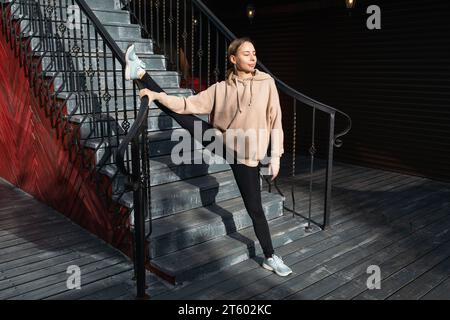 A young attractive woman trains in sportswear on the street, stretches leg muscles with one leg on the railing, basks in the sun standing on one leg w Stock Photo