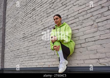 stylish young sportswoman in lime color hoodie and leggings stretching near grey wall outdoors Stock Photo