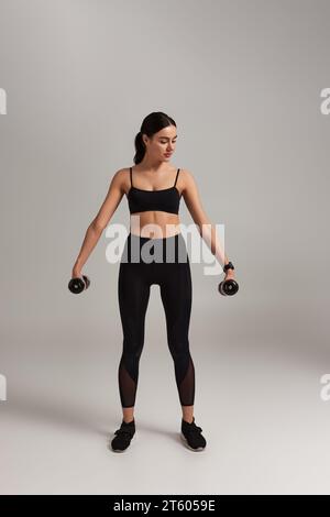 full length of pretty sportswoman in black active wear exercising with dumbbells on grey backdrop Stock Photo