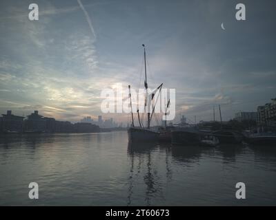 Pearly light of sunrise over River Thames (2). Sailing barge with London docklands in distance, moon. Landscape format. Stock Photo