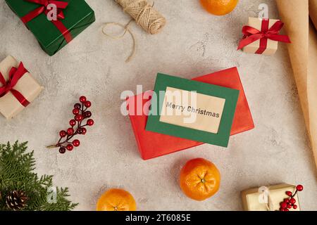 Merry Christmas greeting card near tangerines and decorated gift boxes on grey textured backdrop Stock Photo
