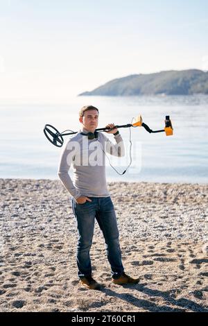 Man with a metal detector on his shoulder stands on the shore and looks ...