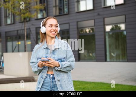 Smiling young woman with headphones holding phone. Joyful lady in denim jacket texts best friend after work to hang out Stock Photo