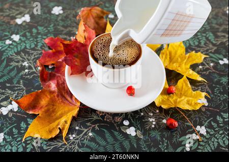 A cup of coffee in autumn mood with autumn colors in the background. Milk flowing from a jug and the orange and red leaves around the cup Stock Photo