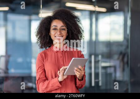 Portrait young successful businesswoman with tablet computer in hands inside office at workplace, female programmer testing new software smiling looking at camera satisfied with achievement results. Stock Photo