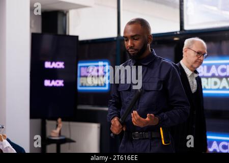 Serious African American man security officer in uniform patrolling shopping mall during Black Friday sales, ensuring crowd control, standing at clothing store entrance preventing shoplifting Stock Photo