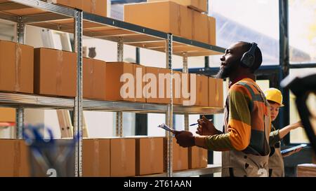 Supervisor inspecting stock logistics with inventory list, looking at merchandise supply chain. Young person examining cardboard boxes for cargo distribution and production, dance moves. Stock Photo