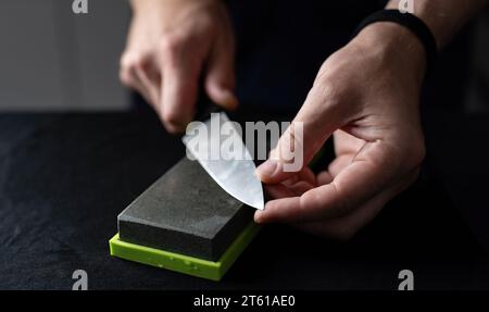 Man Uses Natural Sharpening Whetstone To Sharpen Big Kitchen Knife Against Black Background Stock Photo