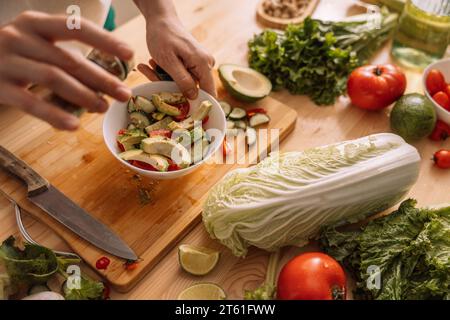 Lady sprinkles spices on vegetarian salad in a white plate on a wooden ...