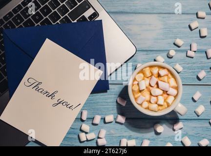 THANK YOU text on card inscription positive quote phrase Greeting card blue envelope with white cup of coffee and marshmallows laptop keyboard on wooden blue table. Workplace holiday. Top view, flat lay minimalist Stock Photo