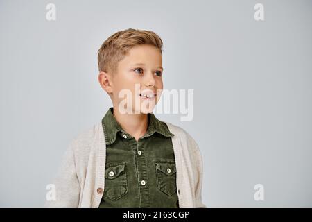 joyful little boy in warm casual cardigan smiling and looking away on gray backdrop, fashion concept Stock Photo