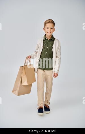 joyous little boy in stylish casual cardigan posing on gray backdrop with present bags, fashion Stock Photo