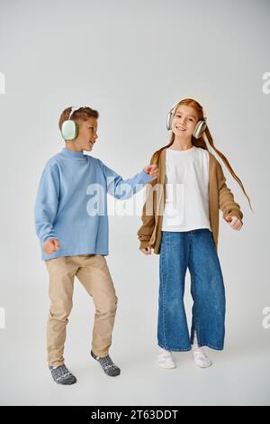 joyous little boy and girl in sweatshirt and cardigan with headphones on gray backdrop, fashion Stock Photo