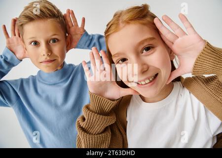 joyous little friends in casual attires smiling at camera with hands near faces, fashion concept Stock Photo