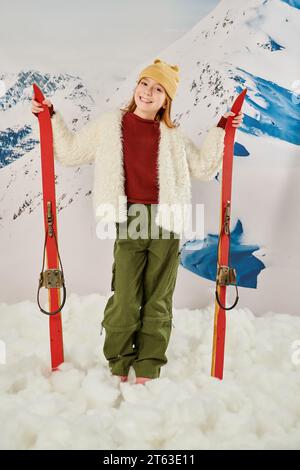 vertical shot of joyous girl in stylish warm clothes holding skis with snowy mountain on backdrop Stock Photo