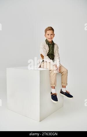 vertical shot of joyous little boy in trendy cardigan sitting on white cube and smiling at camera Stock Photo