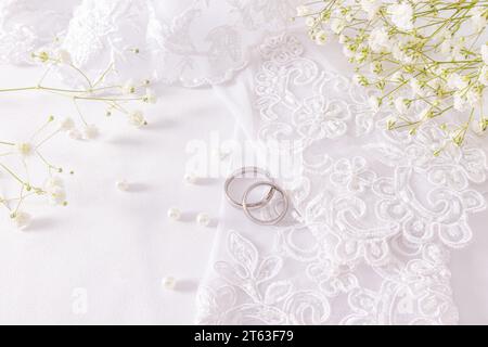 Two white gold wedding rings lie on the bride's white patterned gloves among beads of pearls scattered on white satin. Bride accessories concept Stock Photo
