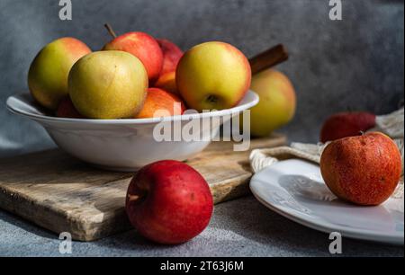 A collection of ripe, colorful apples presented on a rustic wooden board, with a white plate and draped fabric creating a serene kitchen setting Stock Photo