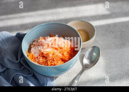 High angle of close-up of a light blue bowl containing freshly grated carrot sprinkled with sugar, placed next to a gray cloth, a small beige bowl wit Stock Photo
