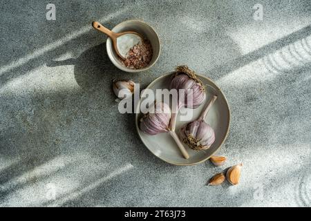 From above of raw garlic bulbs with a bowl of coarse salt on a textured grey surface illuminated by soft sunlight casting shadows Stock Photo