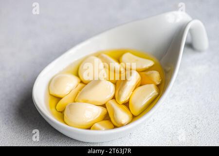 Closeup peeled and baked garlic cloves submerged in oil, showcased in a ceramic spoon against a light gray background Stock Photo