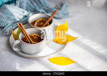 Cups of spiced tea adorned with cinnamon sticks, anise, and dried orange slices, nestled next to vibrant yellow leaves and a folded blue scarf on a so Stock Photo