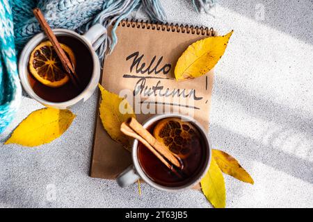 Top view of cups spiced tea with cinnamon sticks and dried orange slices, surrounded by vibrant yellow autumn leaves are juxtaposed against a notebook Stock Photo