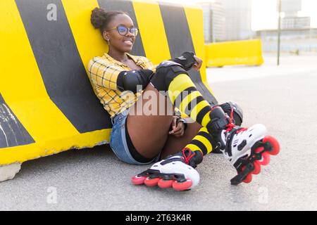 Thoughtful young African American woman in roller skates leaning on barricade while sitting at skate park Stock Photo
