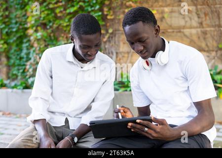 Two African American young men engrossed in a tablet device, sitting outdoors with nature in the backdrop Stock Photo