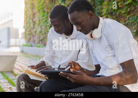 Serious two black young men focusing on their studies one with a book and the other with a tablet seated outdoors near a green wall Stock Photo