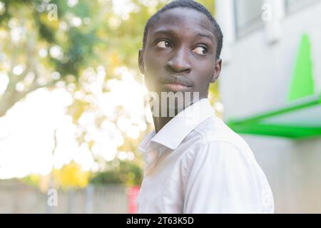 Thoughtful black young man looking away the distance with sunlight filtering through the trees and a city backdrop Stock Photo