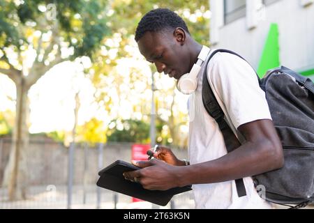 Side view of young man with headphones and a backpack using a tablet on a sunny day with trees in the background Stock Photo