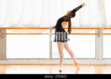 Side view of focused ballet dancer gracefully extends her arm upward balancing on point shoes by the barre in a well lit studio with panoramic windows Stock Photo