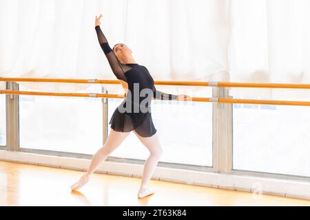 Full body of focused ballet dancer gracefully extends her arm upward balancing on point shoes by the barre in a well lit studio with panoramic windows Stock Photo