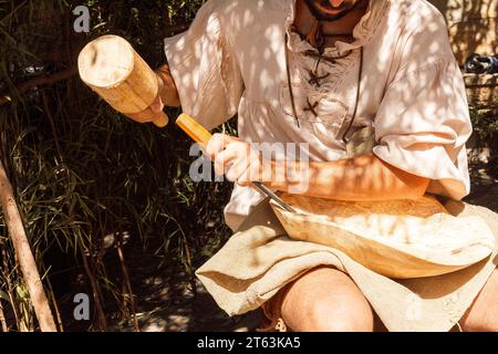 Anonymous medieval craftsman in traditional attire skillfully carves a wooden bowl using hand tools seated amidst greenery Stock Photo