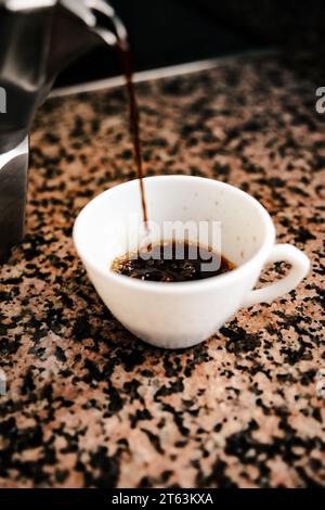 Freshly brewed coffee from a stovetop espresso maker into a white cup set against a modern kitchen backdrop Stock Photo
