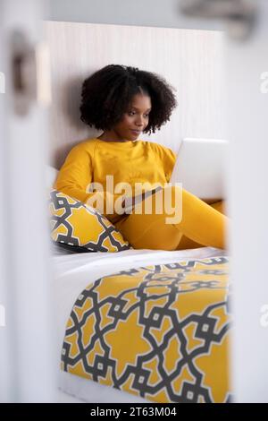 Young African American woman in yellow attire sitting on a bed with patterned cushions engrossed in her laptop in a well lit room Stock Photo