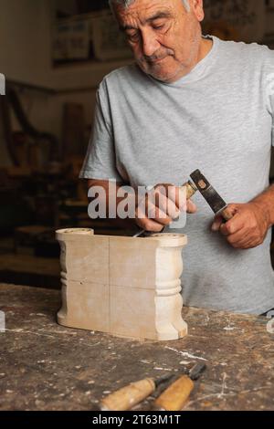 Focused elderly carpenter in Toledo workshop meticulously carving details into a wooden object with a chisel Stock Photo