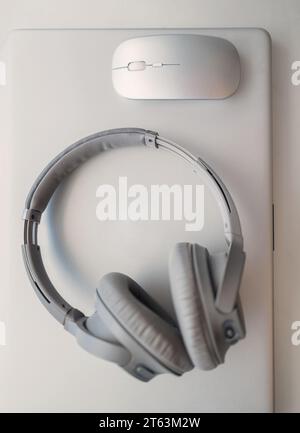 Top view of a pair of silver headphones lying on a white laptop next to a white wireless computer mouse Stock Photo