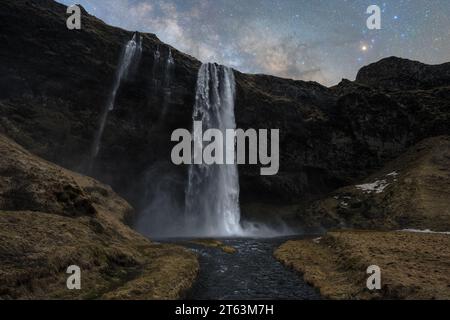 A majestic capture of tall waterfalls cascading down a dark rocky cliff, with a serene river at the base, all set under a starry night sky in Iceland Stock Photo