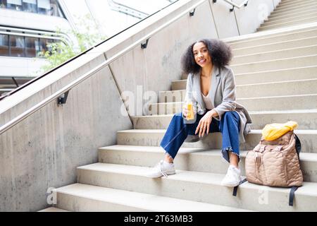 Relaxed afro woman with curly hair sitting on concrete steps outdoors and holding bottle of juice while looking at camera Stock Photo