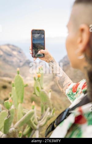Side view of unrecognizable tattooed woman in casual clothes taking selfie with smartphone while standing on background of mountains and cloudy sky in Stock Photo