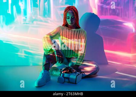 Full body of serious thoughtful young woman with modern VR goggles sitting in glowing lights while exploring cyberspace in studio Stock Photo