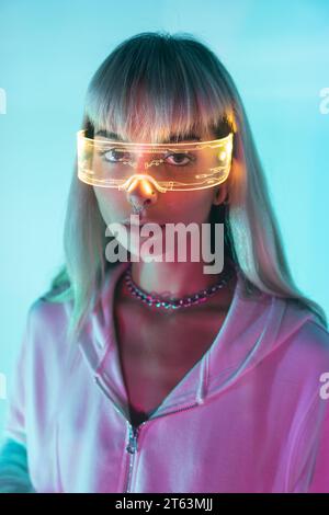 Portrait of confident unemotional young woman wearing LED futuristic VR glasses and looking at camera in blue background Stock Photo