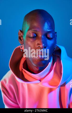 Portrait of depressed bald young man with piercing on face looking down pensively against blue background with illuminated light Stock Photo