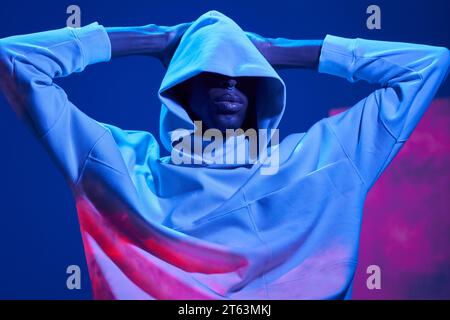 Portrait of thoughtful African American young man dressed in hooded jacket covering eyes with hands on head in studio against blue wall Stock Photo