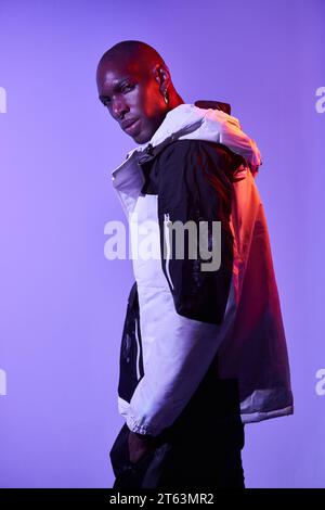 Side view of stylish confident bald young man in casual outfit looking at camera and posing on purple background with glowing lights Stock Photo