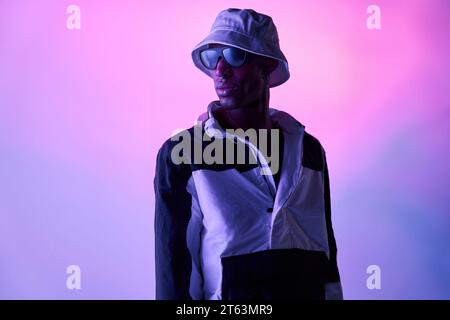 Serious African American young man in stylish outfit sunglasses and hat standing and posing confidently over glowing light in studio while looking awa Stock Photo