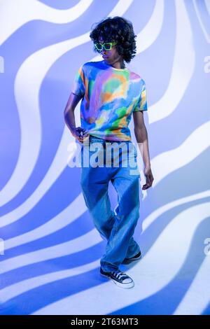 A young man with curly hair strides confidently, showcasing his tie-dye shirt and green sunglasses, complemented by a dynamic wavy blue background Stock Photo